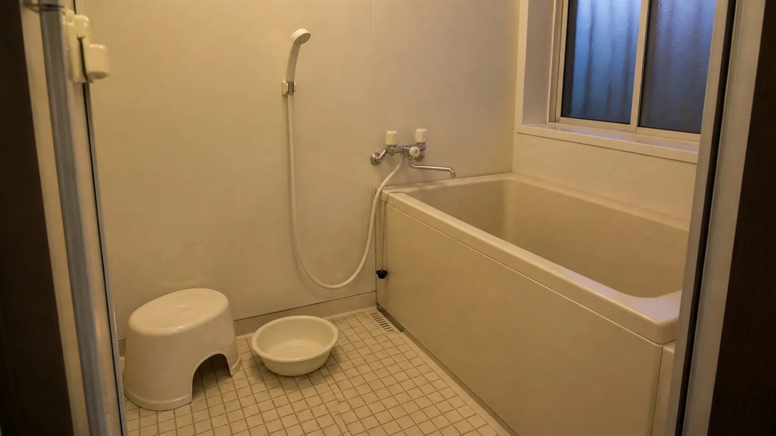 A wide view of a quiet Japanese home bathroom with a wall-mounted hand shower, a low stool, and a short, deep soaking tub
