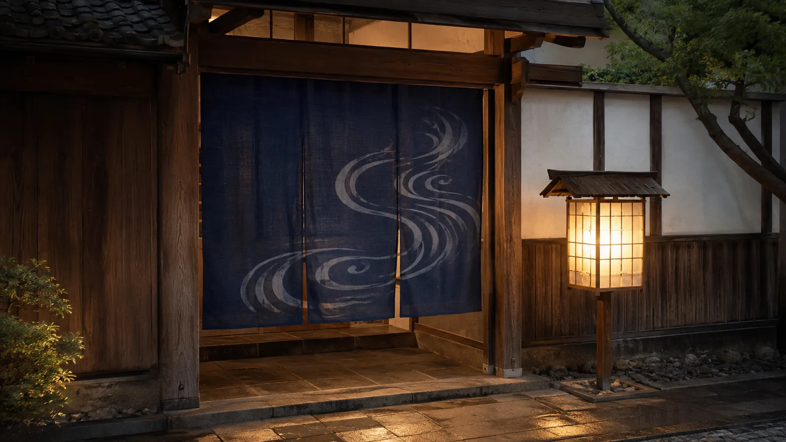 A traditional Japanese bathhouse entrance with an indigo noren curtain and a warmly lit lantern beside it