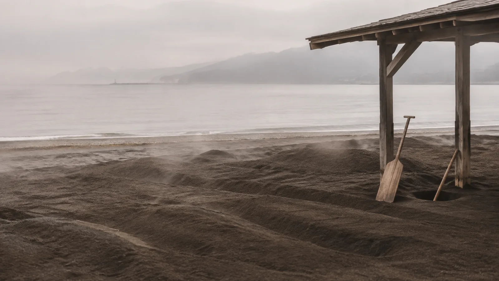 A raked stretch of dark coastal sand under a low wooden shelter with faint steam rising, the sea in the background under soft overcast light